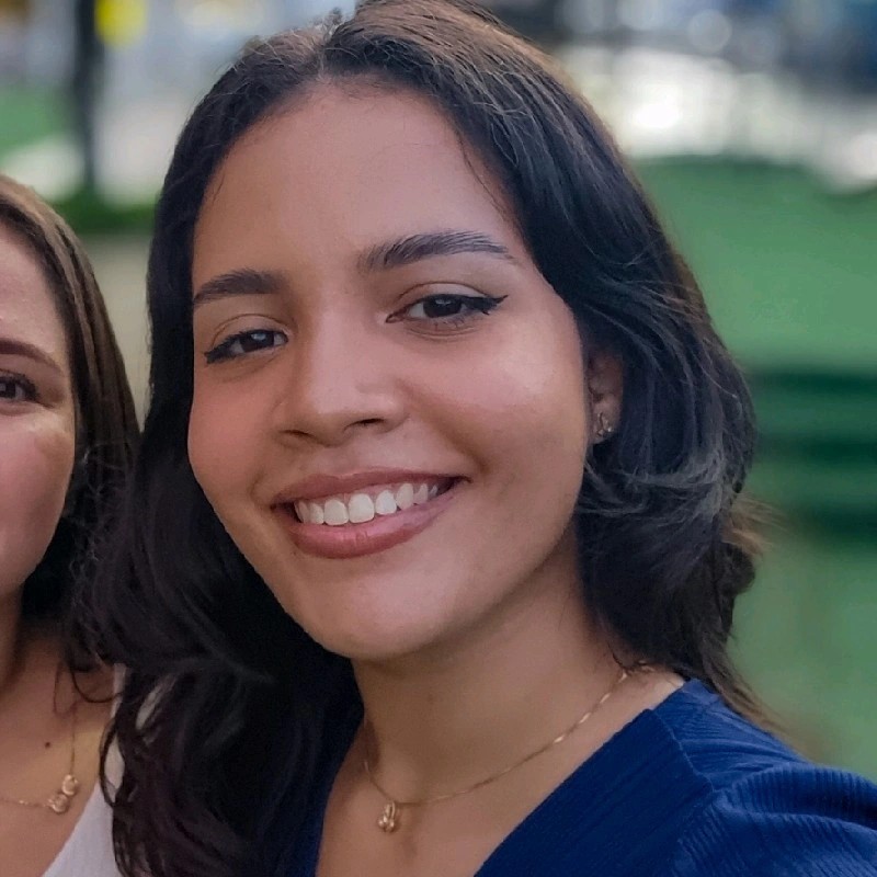 Maria Cibelly de Sousa Castro sorrindo ao ar livre, cabelo castanho curto e blusa azul com fundo verde desfocado