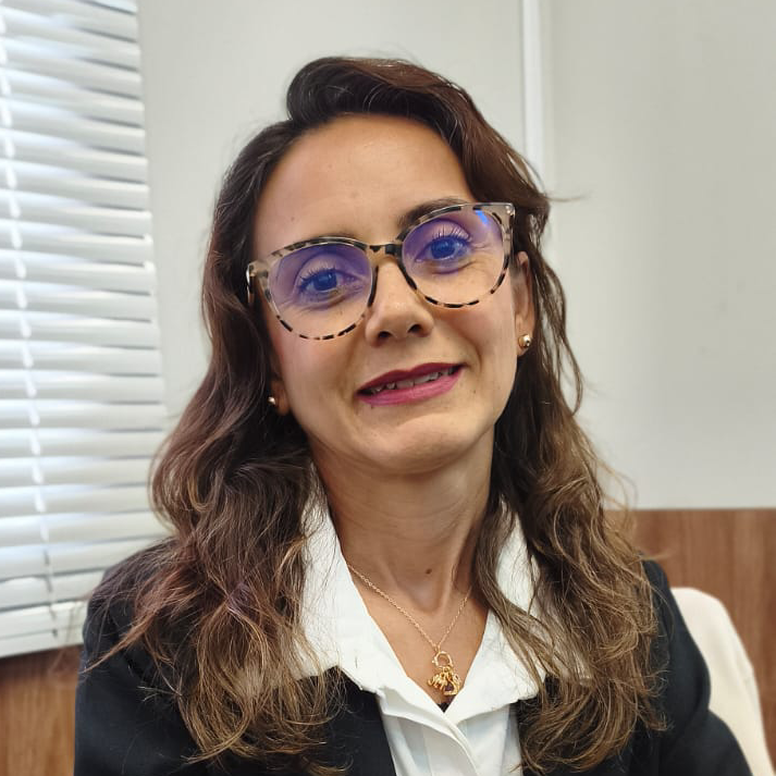 Luiza Manoela Souza da Silva sorrindo, com óculos tartaruga roxos, blazer preto e persianas brancas ao fundo